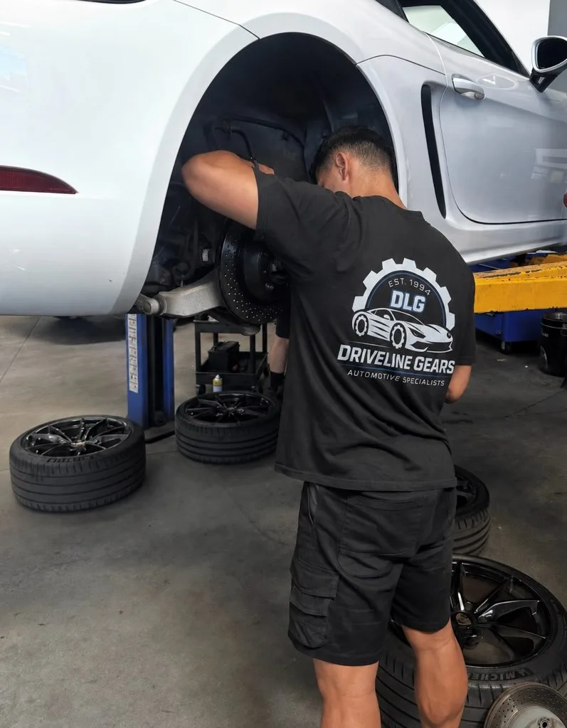 Driveline Gears mechanic in a branded shirt working on the front brake of a white Porsche raised on the hoist, its alloy wheels off and standing on the workshop floor